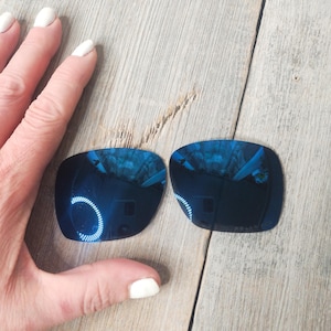 May include: Two blue mirrored replacement lenses for sunglasses. The lenses are rectangular and have a slight curve. They are on a wooden surface.