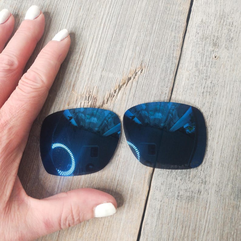 May include: Two blue mirrored replacement lenses for sunglasses. The lenses are rectangular and have a slight curve. They are on a wooden surface.