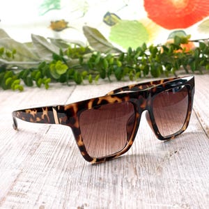 May include: Tortoiseshell sunglasses with brown gradient lenses. The sunglasses have a square frame with a gold accent on the temples. The glasses are set against a white wooden surface.