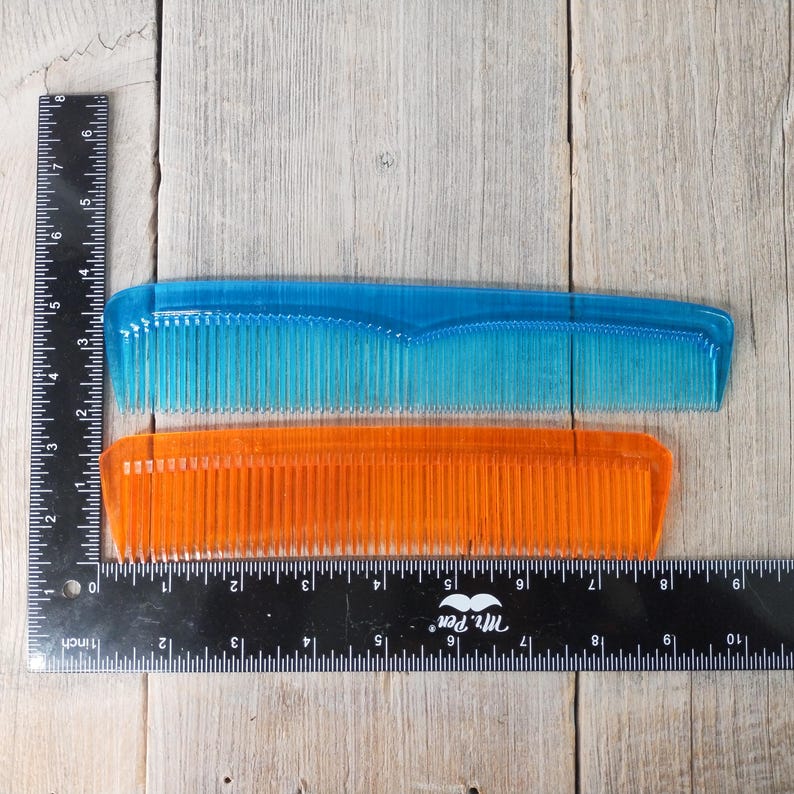May include: Two plastic combs, one blue and one orange, are displayed. The blue comb is approximately 6 inches long, and the orange comb is about 5.5 inches long. A black ruler with inch markings is placed next to the combs.