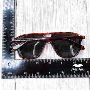 May include: Tortoiseshell sunglasses with dark lenses. The sunglasses are approximately 6 inches wide. The frame has a classic aviator shape. The image is taken from above, showing the sunglasses on a light-colored wooden surface.