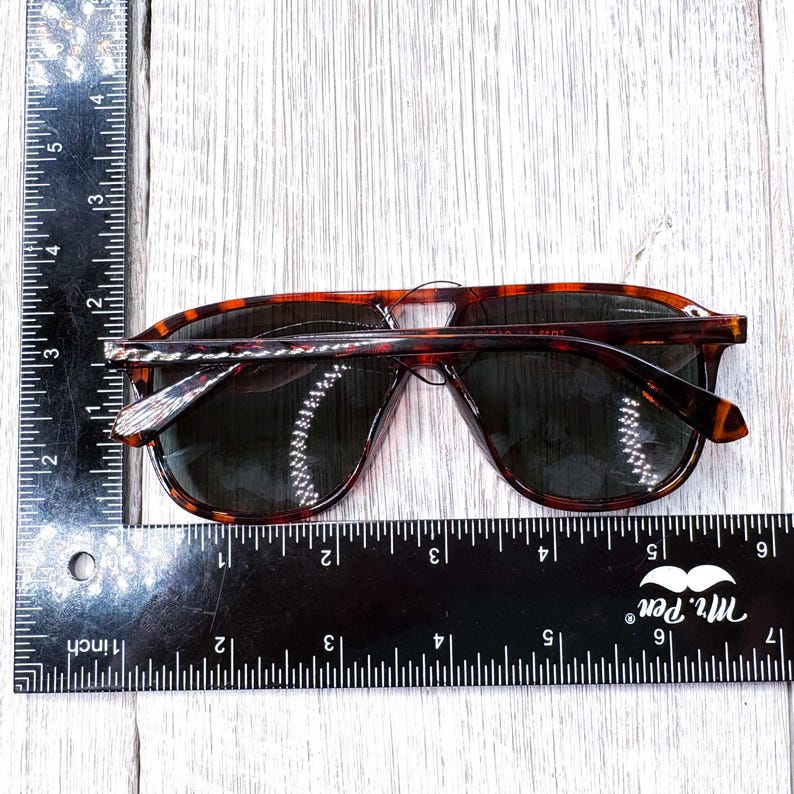 May include: Tortoiseshell sunglasses with dark lenses. The sunglasses are approximately 6 inches wide. The frame has a classic aviator shape. The image is taken from above, showing the sunglasses on a light-colored wooden surface.