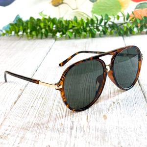 Pode incluir: Óculos de sol estilo aviador com armação de tartaruga em padrão castanho e preto, lentes tingidas de verde escuro e detalhes em metal dourado nas dobradiças e na ponte. A armação tem um formato clássico de gota com ponte dupla.