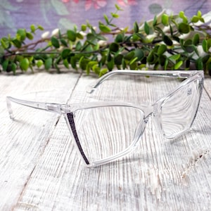 May include: Clear-framed reading glasses with a cat-eye shape. The glasses have a subtle glitter detail on the outer edges of the frames. The lenses have a +1.00 magnification. The glasses are set against a white wooden surface with a floral background.