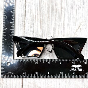 Puede incluir: Gafas de sol negras con dise&ntilde;o de ojo de gato. Se miden con una regla, mostrando una longitud de aproximadamente 15 cm. Las monturas son brillantes y las lentes oscuras.