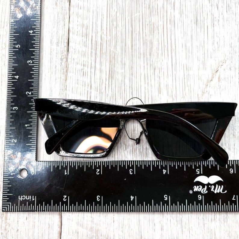 Puede incluir: Gafas de sol negras con dise&ntilde;o de ojo de gato. Se miden con una regla, mostrando una longitud de aproximadamente 15 cm. Las monturas son brillantes y las lentes oscuras.