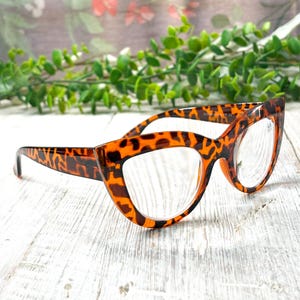 May include: Cat-eye style eyeglasses with a tortoiseshell pattern. The frames are a mix of brown and orange, with clear lenses. The glasses are set against a white wooden surface with greenery in the background.
