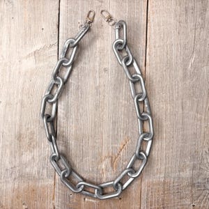 May include: A silver-colored chain with large, rectangular links and two clasp attachments. The chain is displayed against a light brown, wooden background, creating a contrast in textures and tones. The chain is likely for a bag or accessory.