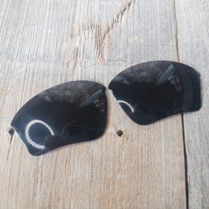 May include: Two black replacement lenses for sunglasses. The lenses are curved and have a reflective surface.