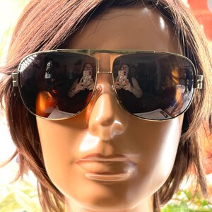 Op de afbeelding: Aviator zonnebril met een goudkleurig metalen montuur, een dubbele brug en donkere, reflecterende glazen. De zonnebril heeft een klassieke druppelvorm met dunne pootjes en ge&iuml;ntegreerde neuspads voor een gestructureerde uitstraling.