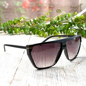 May include: Black sunglasses with a geometric frame and gradient lenses. The sunglasses have a sleek, modern design with a dark frame and lenses that transition from dark at the top to lighter at the bottom. The sunglasses are resting on a white wooden surface.