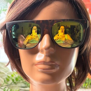 May include: Brown tortoiseshell sunglasses with green tinted lenses are displayed on a mannequin head with brown hair. The lenses reflect a person taking a photo. The sunglasses have the text "UV 400" and "CE" printed on the bottom of the lens.