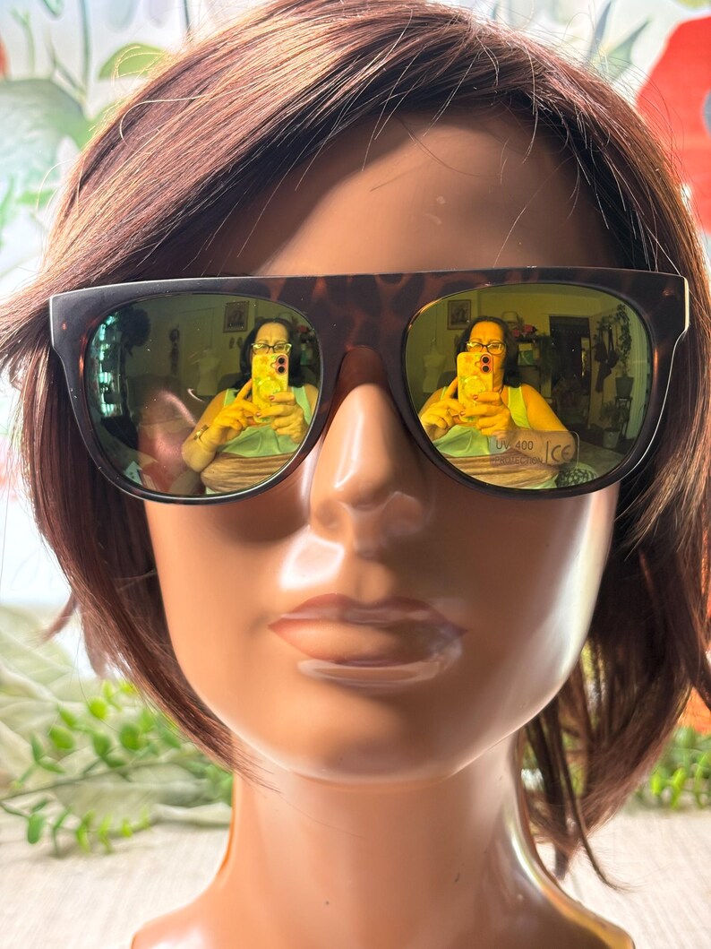 May include: Brown tortoiseshell sunglasses with green tinted lenses are displayed on a mannequin head with brown hair. The lenses reflect a person taking a photo. The sunglasses have the text "UV 400" and "CE" printed on the bottom of the lens.