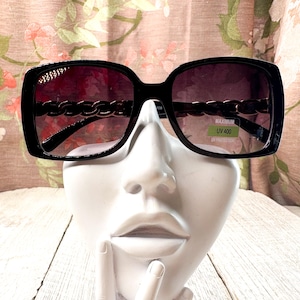 Puede incluir: Gafas de sol cuadradas negras con detalle de cadena en las patillas. Las lentes tienen un degradado de oscuro a claro. Las gafas de sol tienen una pegatina que dice "MAXIMUM UV 400 UV PROTECTION."