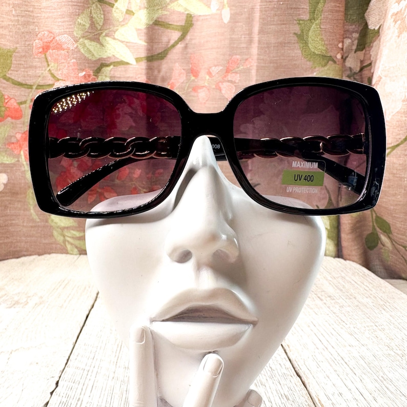 Puede incluir: Gafas de sol cuadradas negras con detalle de cadena en las patillas. Las lentes tienen un degradado de oscuro a claro. Las gafas de sol tienen una pegatina que dice "MAXIMUM UV 400 UV PROTECTION."