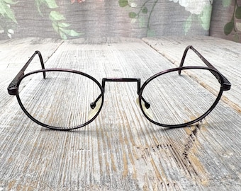 May include: Oval-shaped eyeglasses with a dark burgundy frame. The glasses feature a bridge and thin arms. The clear lenses are set against a weathered wooden surface. The frame's colour is a deep, rich burgundy.