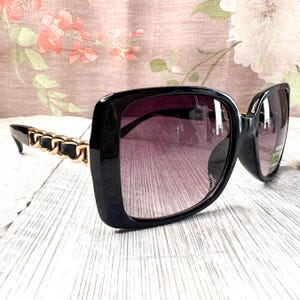Puede incluir: Gafas de sol cuadradas negras con lentes degradadas. Las patillas presentan un dise&ntilde;o de cadena dorada. Las gafas de sol est&aacute;n sobre un fondo floral y una superficie de madera blanca.