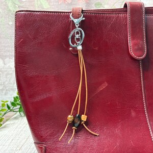 May include: A burgundy leather tote bag with a matching handle. A silver-toned keyring with a leather tassel and wooden beads hangs from the bag. The bag has visible stitching and a smooth texture.