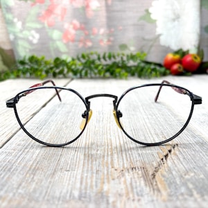 May include: Black oval-shaped eyeglasses with thin frames and a bridge. The temples have a red and black pattern. The lenses are clear. The glasses are set against a blurred background of flowers and greenery.