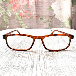 May include: Brown tortoiseshell eyeglasses with a rectangular frame. The glasses have clear lenses and are set against a white wooden surface with a floral patterned curtain in the background.