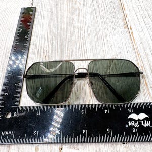 Pode incluir: &Oacute;culos de sol estilo aviador com lentes verde escuro e arma&ccedil;&atilde;o met&aacute;lica fina. Os &oacute;culos est&atilde;o sobre uma superf&iacute;cie de madeira clara, com uma r&eacute;gua pr&oacute;xima para escala. As lentes t&ecirc;m aproximadamente 5 cm de largura.