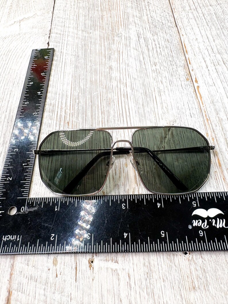 Pode incluir: &Oacute;culos de sol estilo aviador com lentes verde escuro e arma&ccedil;&atilde;o met&aacute;lica fina. Os &oacute;culos est&atilde;o sobre uma superf&iacute;cie de madeira clara, com uma r&eacute;gua pr&oacute;xima para escala. As lentes t&ecirc;m aproximadamente 5 cm de largura.