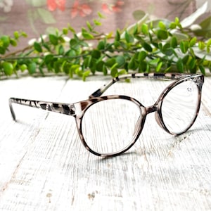 May include: A pair of round-framed reading glasses with a tortoiseshell pattern on the arms and frame. The lenses have a +2.75 magnification. The glasses are set against a white wooden surface with green foliage and a floral background.