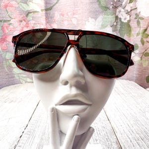 May include: Brown tortoiseshell sunglasses with dark lenses. The sunglasses have a double bridge and a classic aviator shape. The frames are a warm brown color with a mottled pattern. The sunglasses are displayed on a white bust.