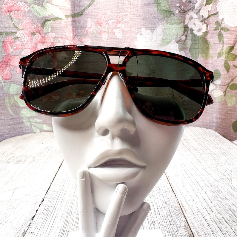 May include: Brown tortoiseshell sunglasses with dark lenses. The sunglasses have a double bridge and a classic aviator shape. The frames are a warm brown color with a mottled pattern. The sunglasses are displayed on a white bust.
