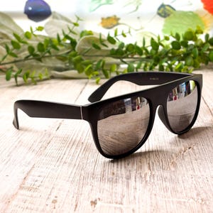 May include: Black sunglasses with silver mirrored lenses. The sunglasses have a thick, rectangular frame and are set against a light-colored wooden surface. The lenses reflect the surrounding environment.