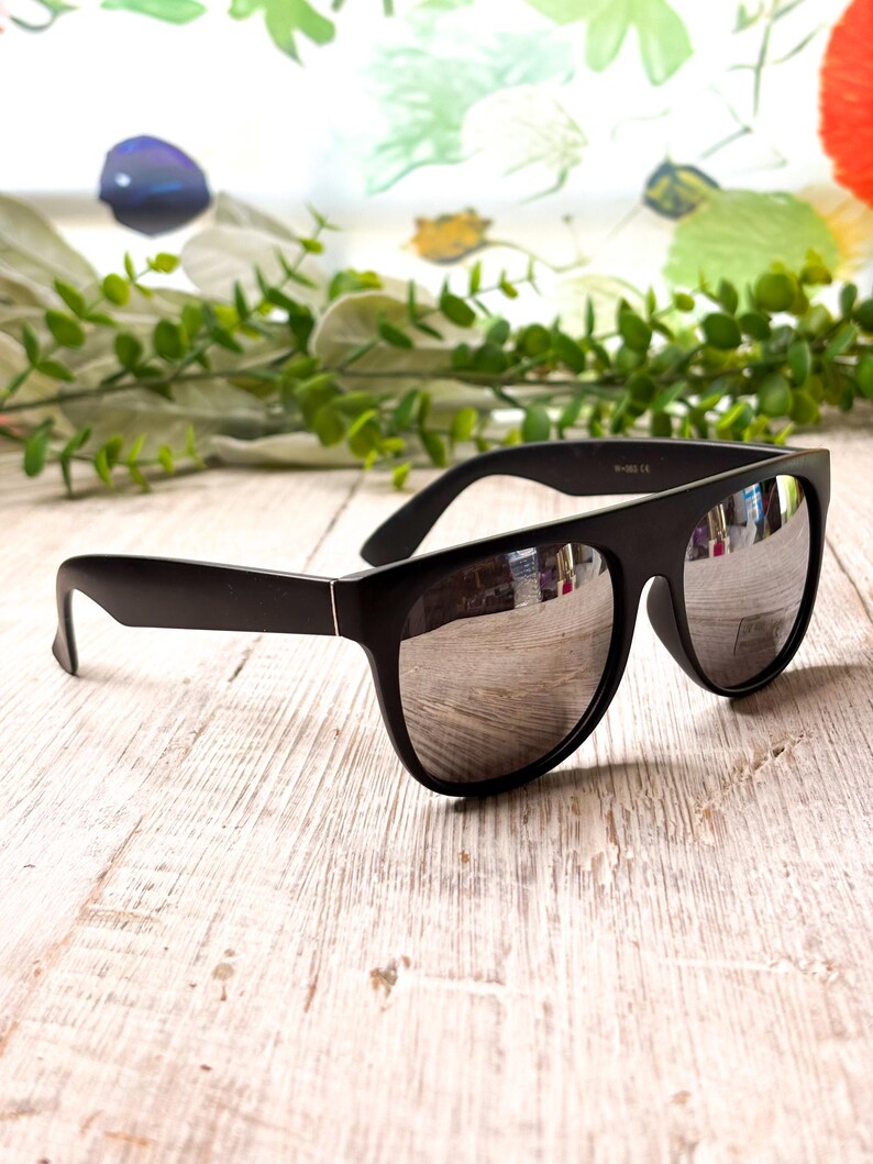 May include: Black sunglasses with silver mirrored lenses. The sunglasses have a thick, rectangular frame and are set against a light-colored wooden surface. The lenses reflect the surrounding environment.