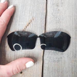 Op de afbeelding: Een paar zwarte vervangende glazen voor zonnebrillen. De glazen zijn gebogen en hebben een zilveren ring rond de rand.