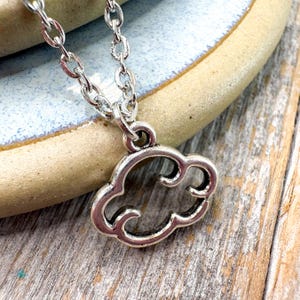 May include: Silver-toned cloud pendant necklace. The pendant is a stylized cloud shape with a cutout design. The necklace chain is a delicate link style. The necklace rests on a light blue and tan ceramic surface and a wooden surface.