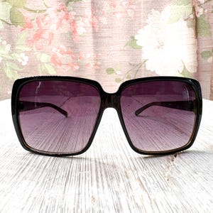 以下が含まれることがあります： 黒いスクエアフレームのサングラス、紫色のグラデーションレンズ付き。 サングラスは太いフレームとクラシックなデザインです。 レンズは上部で濃い色合いから下部で明るい色合いに変化します。