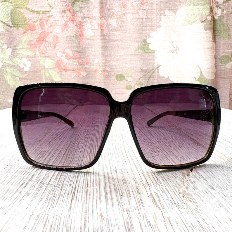 以下が含まれることがあります： 黒いスクエアフレームのサングラス、紫色のグラデーションレンズ付き。 サングラスは太いフレームとクラシックなデザインです。 レンズは上部で濃い色合いから下部で明るい色合いに変化します。