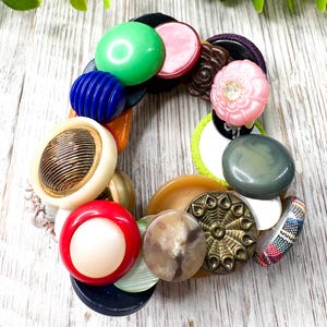 May include: A colorful bracelet made of various vintage buttons. The buttons are in shades of green, pink, blue, red, and brown. Some buttons have intricate designs, while others are smooth and solid. The bracelet is on a light wood surface.