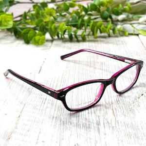 Puede incluir: Gafas negras y rosas con montura rectangular. Las gafas tienen interior y patillas rosas, con el texto "DEMO LENS ONLY" visible en la lente. Las gafas están sobre una superficie de madera blanca con follaje verde en el fondo.
