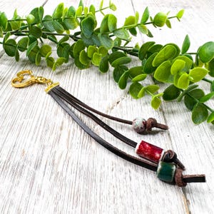 May include: A brown leather keychain with a gold-colored clasp. The keychain features three leather straps, each adorned with a decorative bead in red, green, and speckled white. The background includes green foliage.