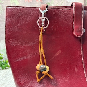 May include: A close-up of a red leather tote bag with a decorative keychain. The keychain features a silver clasp, a leather tassel, and three wooden beads in shades of brown and tan.