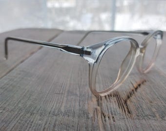 May include: Clear plastic framed spectacles with round lenses. The glasses are on a wooden surface.