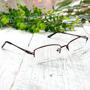 May include: A pair of Christie Brinkley eyeglasses with a burgundy frame and a half-rim design. The glasses have a rectangular shape and are set against a white wooden surface with green foliage in the background.