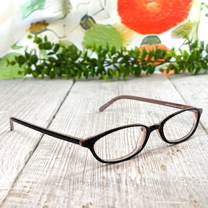 May include: Eyeglasses featuring a black frame front with a contrasting light pink interior and matching light pink temples. The lenses are clear and oval-shaped, with a thin bridge connecting the two rims. The design is simple and functional.