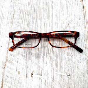 Puede incluir: Una taza de cer&aacute;mica blanca con un asa negra y el texto "But First, Coffee" en negrita negra. La taza se encuentra sobre un fondo borroso, sugiriendo un ambiente acogedor.