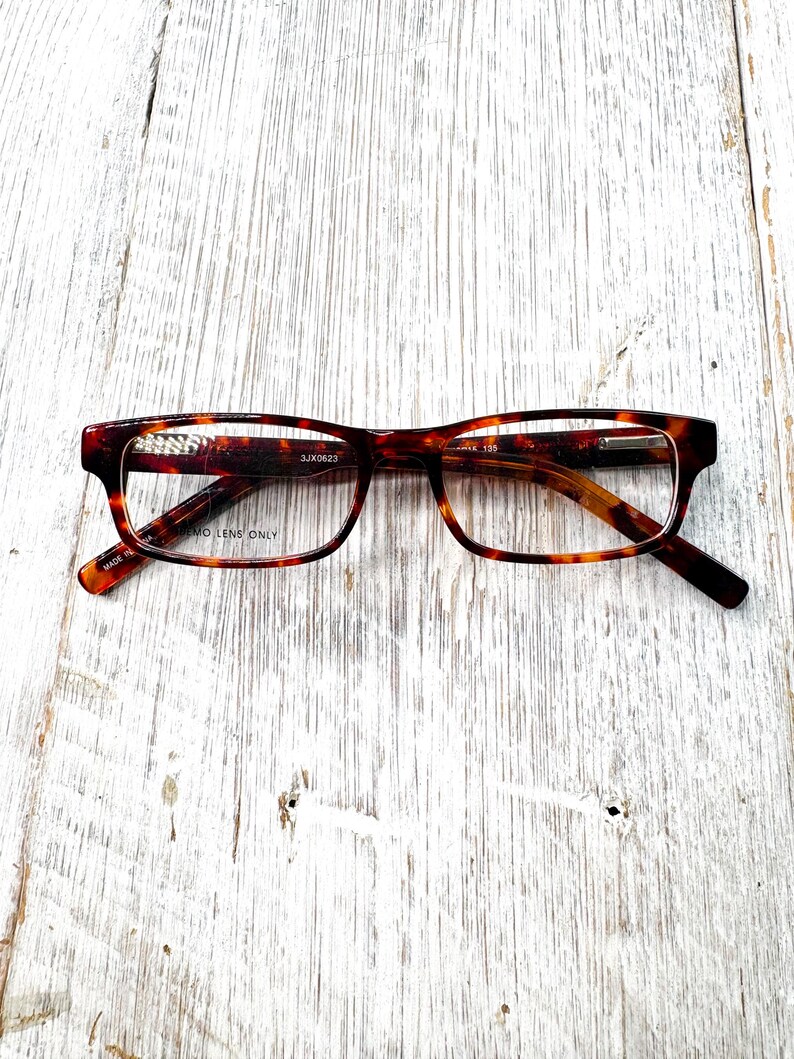 Puede incluir: Una taza de cer&aacute;mica blanca con un asa negra y el texto "But First, Coffee" en negrita negra. La taza se encuentra sobre un fondo borroso, sugiriendo un ambiente acogedor.