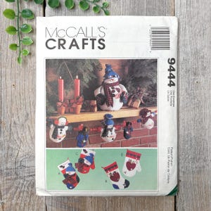 Op de afbeelding: Een vintage McCall's Crafts naaipatroon voor het maken van kerstversiering. Het patroon bevat instructies voor het maken van een sneeuwpop, wanten en een krans. Het patroonnummer is 9444.