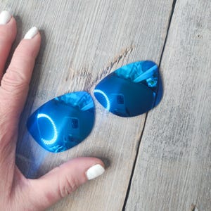 May include: Two blue mirrored replacement lenses for sunglasses. The lenses are curved and have a reflective surface.