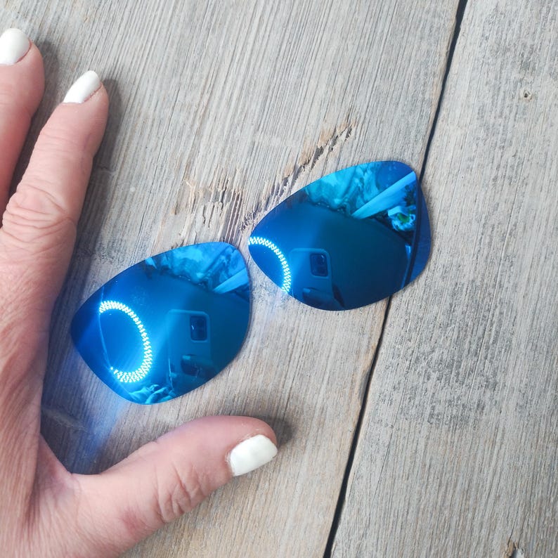 May include: Two blue mirrored replacement lenses for sunglasses. The lenses are curved and have a reflective surface.