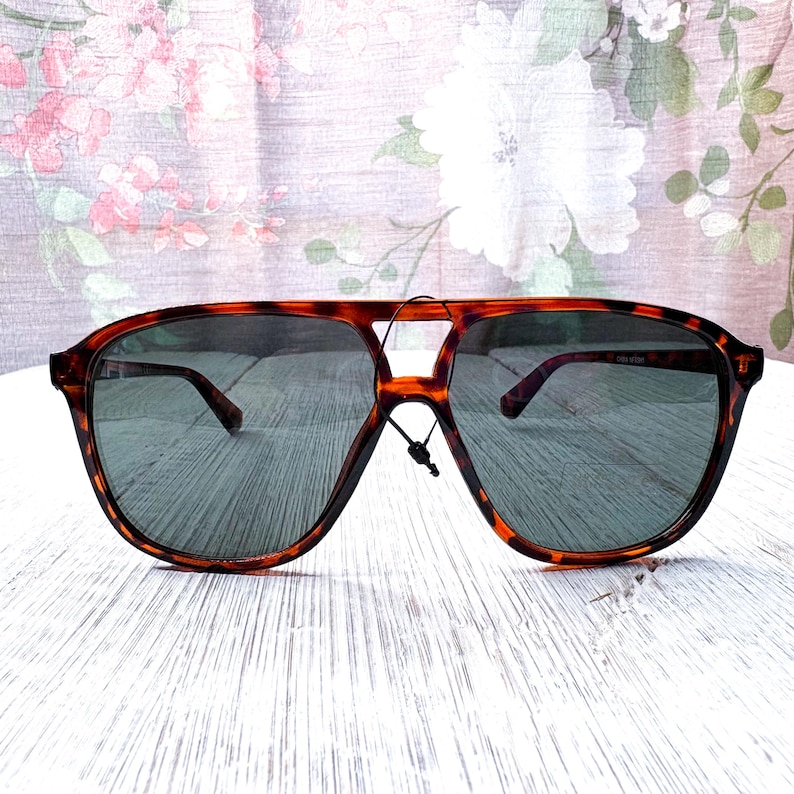 May include: Tortoiseshell-framed sunglasses with dark lenses. The sunglasses have a classic aviator shape with a double bridge and a keyhole nose bridge. The frames are a warm brown color with a mottled pattern. The lenses are a dark gray color.