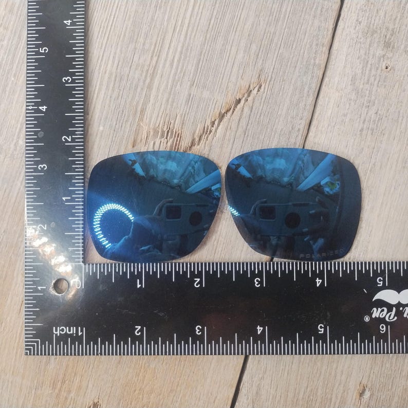 May include: Two blue replacement lenses for sunglasses. The lenses are rectangular and have a slight curve. They are measured against a ruler in inches.