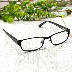 May include: Black rectangular eyeglasses with clear demo lenses. The frames are a matte black and feature a thin, modern design. The text "DEMO LENS ONLY" is visible on one lens. The glasses are set against a light wood-grain surface with greenery in the background.